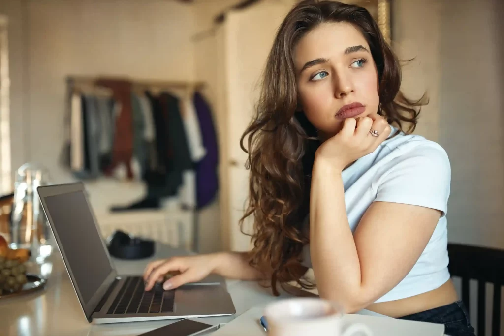 Mulher sentada à mesa usando um notebook, apoiando o rosto na mão e olhando pensativa para a tela, em um ambiente doméstico