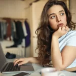 Mulher sentada à mesa usando um notebook, apoiando o rosto na mão e olhando pensativa para a tela, em um ambiente doméstico
