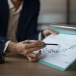 Pessoa sentada à mesa analisando um documento impresso com linha do tempo e indicadores de crescimento, apontando com uma caneta para um gráfico que sobe
