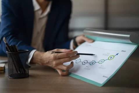 Pessoa sentada à mesa analisando um documento impresso com linha do tempo e indicadores de crescimento, apontando com uma caneta para um gráfico que sobe