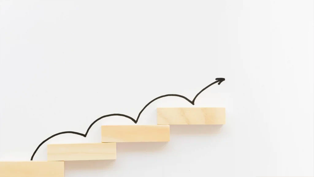 Degraus de madeira dispostos em sequência crescente sobre fundo branco, com uma seta desenhada indicando avanço passo a passo