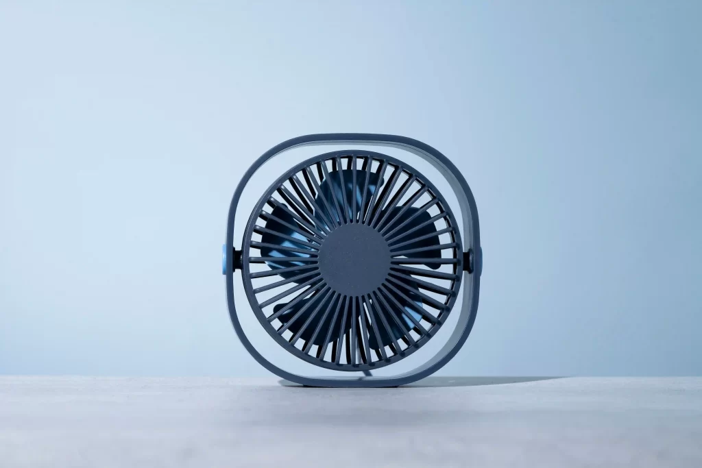 Ventilador portátil de mesa na cor azul, visto de frente, apoiado sobre uma superfície clara, com fundo azul claro e design compacto