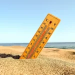 Termômetro analógico cravado na areia da praia indicando temperatura elevada, com o mar e o céu azul ao fundo em um dia ensolarado