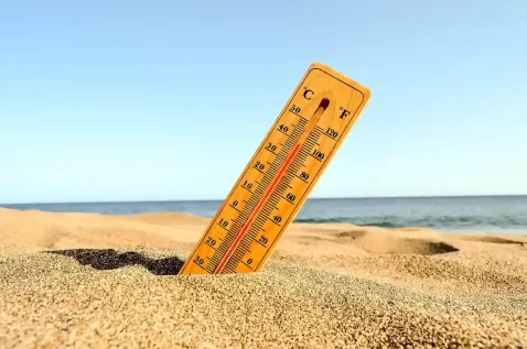 Termômetro analógico cravado na areia da praia indicando temperatura elevada, com o mar e o céu azul ao fundo em um dia ensolarado
