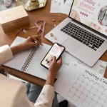 Pessoa sentada à mesa escreve em um caderno enquanto segura um celular. Na frente dela há um notebook aberto com uma página de planejamento digital