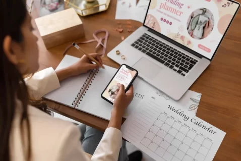 Pessoa sentada à mesa escreve em um caderno enquanto segura um celular. Na frente dela há um notebook aberto com uma página de planejamento digital