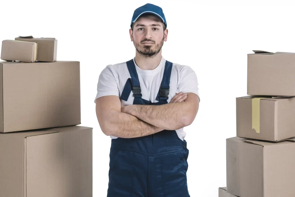 Homem usando macacão azul e boné, com braços cruzados, em pé entre pilhas de caixas de papelão, sobre fundo branco