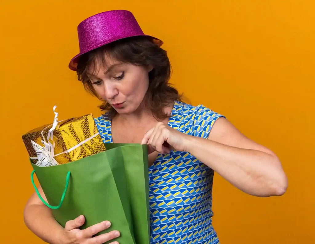 Mulher adulta usando chapéu rosa brilhante e vestido estampado olha com curiosidade para dentro de uma sacola verde de compras