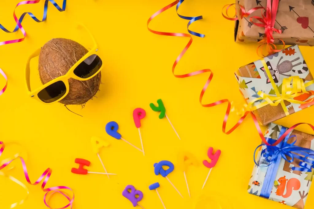 Composição colorida sobre fundo amarelo com um coco usando óculos escuros, caixas de presente embrulhadas com fitas coloridas, serpentinas e letras decorativas formando a palavra “party”