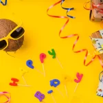 Composição colorida sobre fundo amarelo com um coco usando óculos escuros, caixas de presente embrulhadas com fitas coloridas, serpentinas e letras decorativas formando a palavra “party”