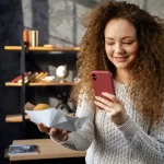 Mulher com cabelos cacheados, em um ambiente interno, segura um celular com uma mão enquanto observa um alimento embalado na outra