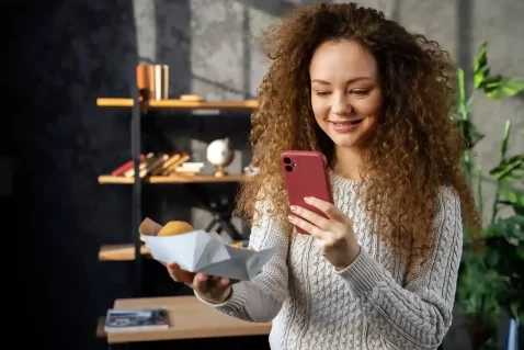 Mulher com cabelos cacheados, em um ambiente interno, segura um celular com uma mão enquanto observa um alimento embalado na outra