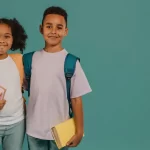 Crianças em pé, lado a lado, usando mochilas escolares nas costas. A menina segura um caderno rosa e o menino segura um livro amarelo