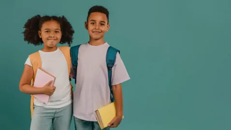 Crianças em pé, lado a lado, usando mochilas escolares nas costas. A menina segura um caderno rosa e o menino segura um livro amarelo