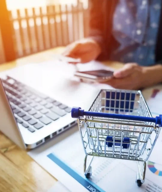 Mini carrinho de compras sobre gráficos e notebook enquanto pessoa usa celular, simbolizando análise de demanda Mercado Livre baseada em dados de vendas