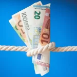 Pessoa segura notas de dinheiro presas por um nó em uma corda contra fundo azul, representando a pressão financeira e os desafios de como competir no marketplace