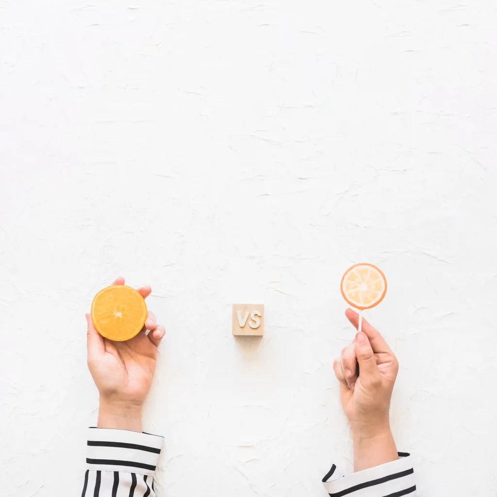 Mãos comparando duas formas de laranja, uma inteira e outra em fatia, com símbolo de versus ao centro, representando comparação de opções antes da compra online