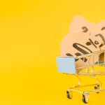 Carrinho de compras em fundo amarelo contendo papéis com anotações de valores e porcentagens, simbolizando o cálculo e a composição dos custos de produtos no e-commerce