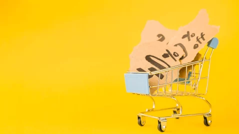 Carrinho de compras em fundo amarelo contendo papéis com anotações de valores e porcentagens, simbolizando o cálculo e a composição dos custos de produtos no e-commerce
