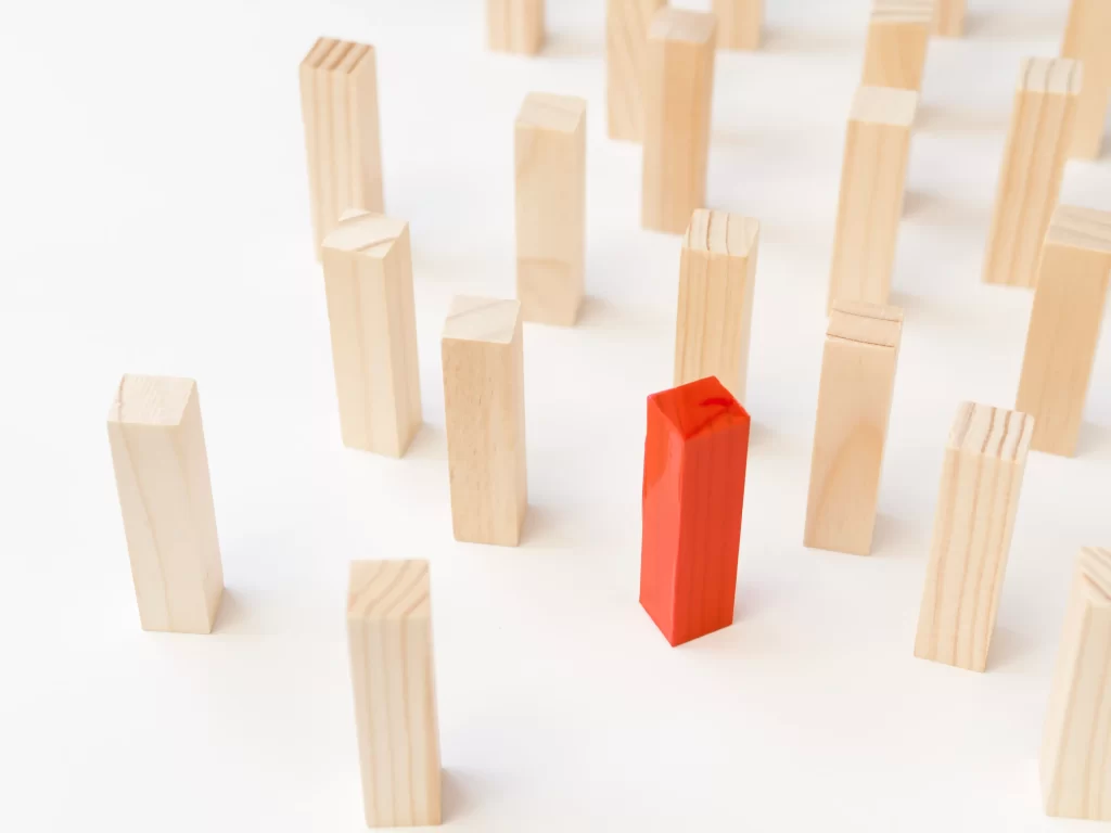 Blocos de madeira claros posicionados em pé sobre fundo branco, com um único bloco vermelho no centro se destacando entre os demais