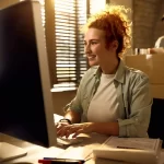 Mulher trabalhando em frente a um computador em um ambiente de operação logística, com caixas ao fundo, representando a rotina de controle, organização e eficiência operacional no e-commerce