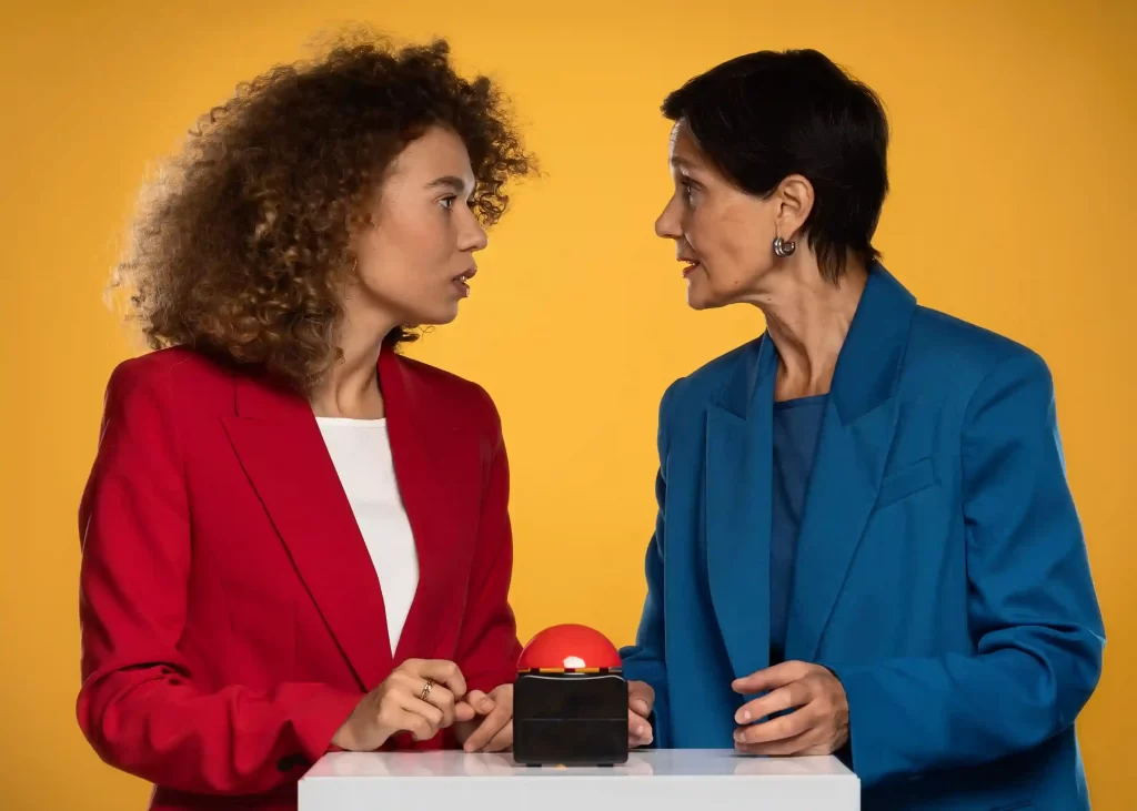 Duas mulheres em fundo amarelo analisando botão vermelho sobre mesa, simbolizando tomada de decisão estratégica na estratégia de preços Mercado Livre