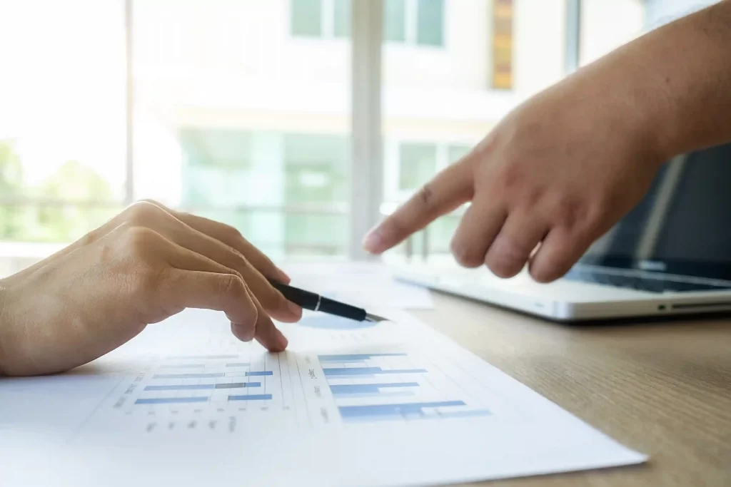 Pessoa apontando para gráficos financeiros impressos sobre mesa com notebook ao fundo, representando análise de dados ligada à formalização no Mercado Livre