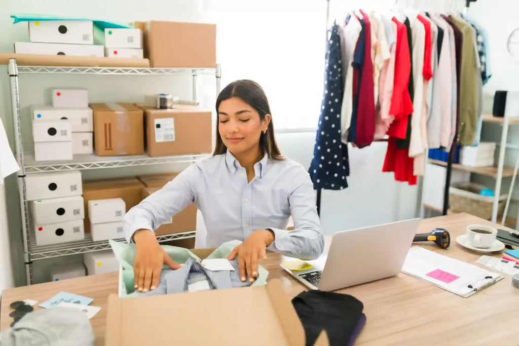 Mulher organizando e embalando roupas em mesa com caixas e arara ao fundo, representando curadoria de produtos em marketplace vertical.