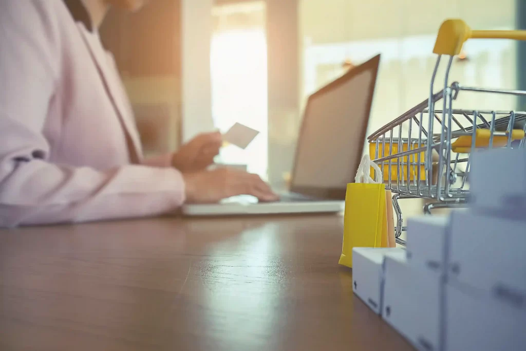 Pessoa usando notebook para comprar online, com mini carrinho de compras e caixas sobre a mesa, ilustrando a dinâmica dos marketplaces no varejo