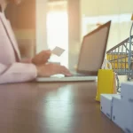 Pessoa usando notebook para comprar online, com mini carrinho de compras e caixas sobre a mesa, ilustrando a dinâmica dos marketplaces no varejo