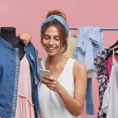 Mulher analisando roupas e usando celular em ambiente de loja, representando o crescimento do mercado de moda e-commerce em 2026.
