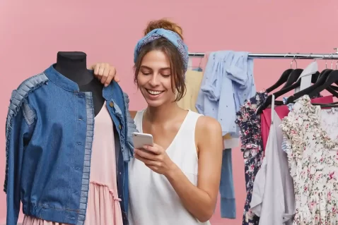 Mulher analisando roupas e usando celular em ambiente de loja, representando o crescimento do mercado de moda e-commerce em 2026.