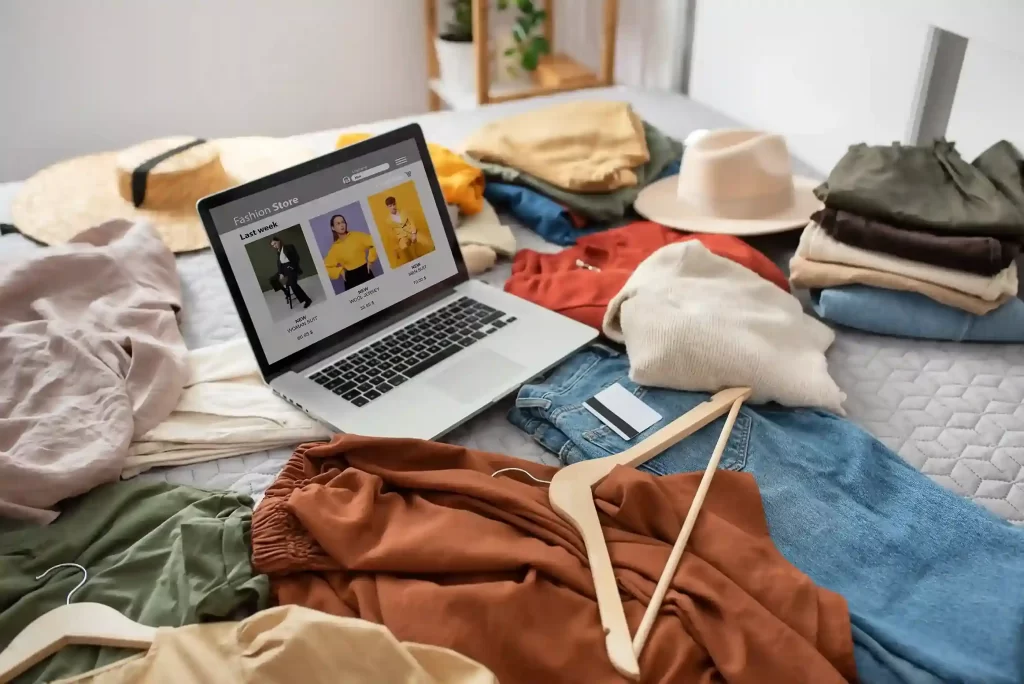 Notebook com loja virtual de roupas aberto sobre a cama cercado por peças de vestuário, simbolizando estratégia no mercado de moda e-commerce