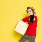 Criança segurando um smartphone e uma caixa de encomenda, em frente a um fundo amarelo