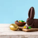 Coelho de chocolate ao lado de ovos de Páscoa e confeitos coloridos sobre uma mesa de madeira clara, com fundo azul, representando produtos típicos da Páscoa