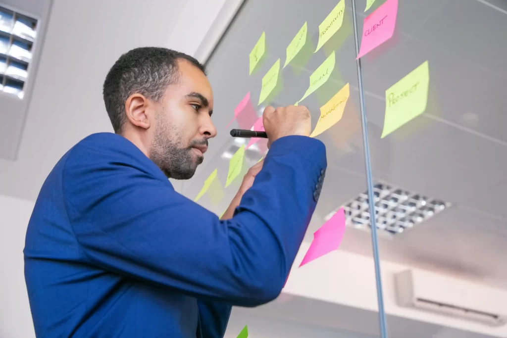 Homem de blazer azul escrevendo em post-its coloridos colados em uma parede de vidro, organizando ideias e tarefas para planejamento e-commerce no pós-Carnaval