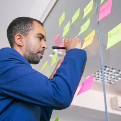 Homem de blazer azul escrevendo em post-its coloridos colados em uma parede de vidro, organizando ideias e tarefas para planejamento e-commerce no pós-Carnaval