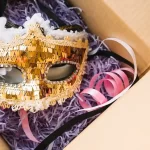 Máscara dourada de Carnaval com detalhes em lantejoulas e fitas coloridas dentro de uma caixa aberta, representando produtos sazonais de giro rápido prontos para venda