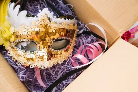 Máscara dourada de Carnaval com detalhes em lantejoulas e fitas coloridas dentro de uma caixa aberta, representando produtos sazonais de giro rápido prontos para venda