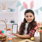 Menina com orelhas de coelho segurando celular ao lado de cesta com ovos coloridos e flores, simbolizando vendas na Páscoa no ambiente digital