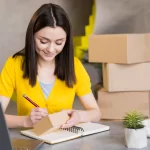 Mulher sentada à mesa anota informações em um caderno enquanto segura uma pequena caixa de papelão, com notebook aberto ao lado e caixas empilhadas ao fundo, ilustrando a rotina de quem vende no Mercado Livre