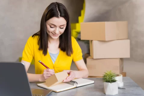 Mulher sentada à mesa anota informações em um caderno enquanto segura uma pequena caixa de papelão, com notebook aberto ao lado e caixas empilhadas ao fundo, ilustrando a rotina de quem vende no Mercado Livre