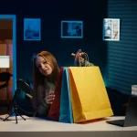 Criadora de conteúdo mostrando sacolas de compras durante transmissão online com computador e microfone, representando o comportamento de compras online à noite