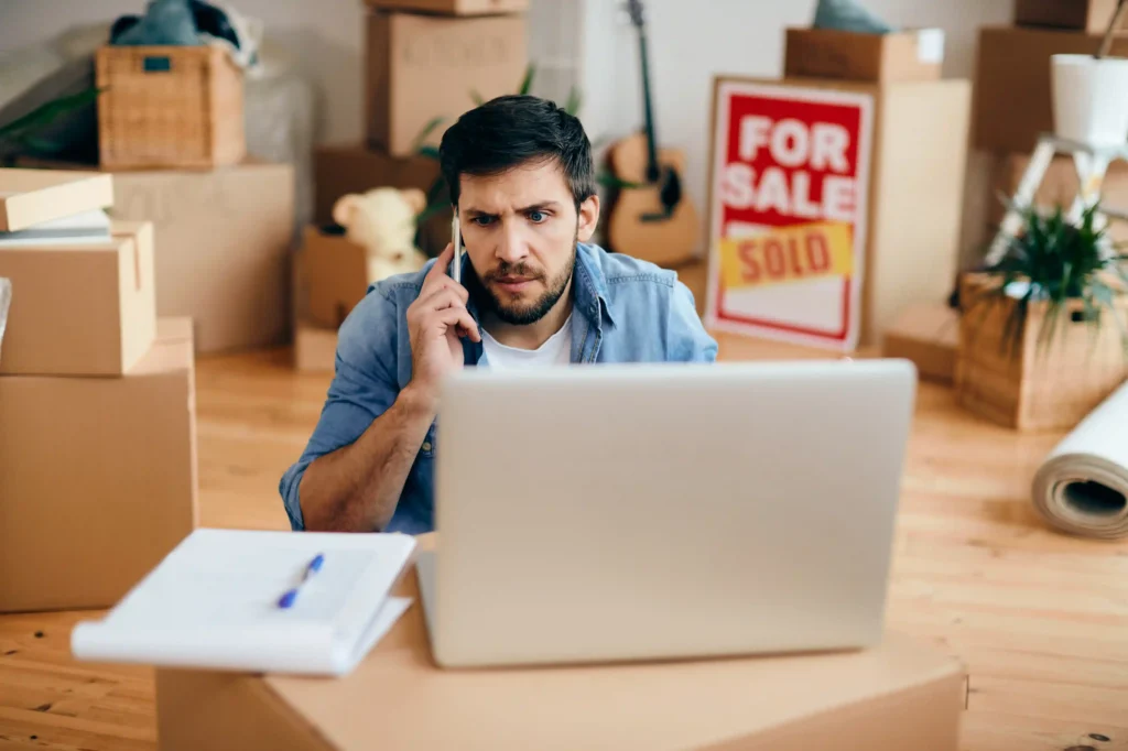 Homem analisando laptop com expressão de preocupação em meio a caixas e anúncio de venda, representando dificuldade em analisar concorrentes no e-commerce