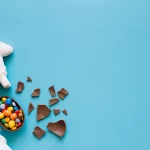 Ovo de chocolate quebrado com doces coloridos ao lado de decoração de Páscoa sobre fundo azul, representando mercado e concorrência na Páscoa 2026