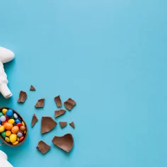 Ovo de chocolate quebrado com doces coloridos ao lado de decoração de Páscoa sobre fundo azul, representando mercado e concorrência na Páscoa 2026
