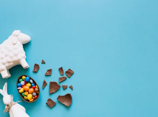 Ovo de chocolate quebrado com doces coloridos ao lado de decoração de Páscoa sobre fundo azul, representando mercado e concorrência na Páscoa 2026