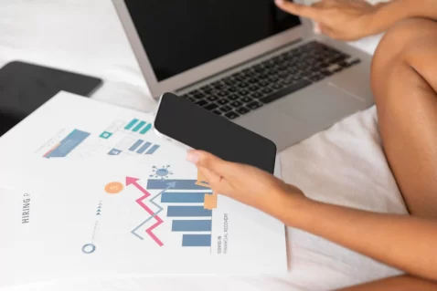 Pessoa analisando gráficos de desempenho no laptop e smartphone para avaliar dados de visitas e tendências de vendas online