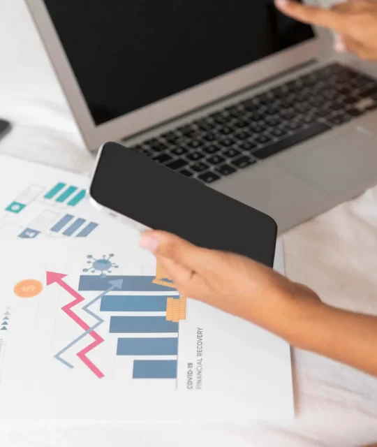 Pessoa analisando gráficos de desempenho no laptop e smartphone para avaliar dados de visitas e tendências de vendas online