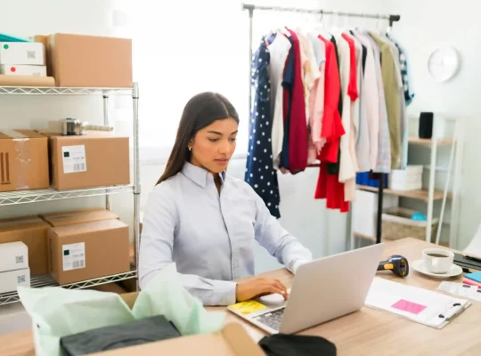 Empreendedora trabalhando em laptop em pequeno estoque com roupas e caixas de envio ao fundo, representando rotina de gestão de e-commerce